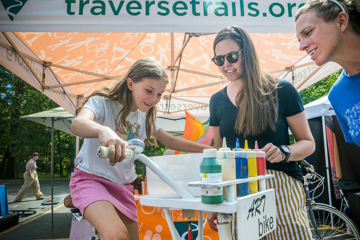 Young girl pedals an art bike while adults look at her under a TentCraft tent.